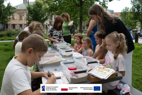 Grupa osób podczas warsztatów archeologicznych siedząca przy stole piknikowym rozstawionym na zielonej trawie na placu Wolności. W tle budynek archiwum oraz przypadkowi przechodnie. 