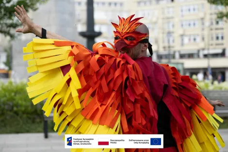 Uśmiechnięty mężczyzna ubrany w strój mitycznego ptaka – feniksa. Na twarzy ma maskę imitującą dziób ptaka, na ramionach pelerynę imitującą pióra ptaka w kolorach intensywnej czerwieni i odcieniach żółtego. Mężczyzna ma rozłożone ręce udając, że rozkłada „skrzydła”. 