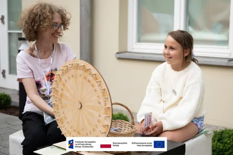 Kobieta w średnim wieku w okularach z krótkimi kręconymi włosami uśmiechająca się do dziewczynki siedzącej po jej prawej. Dziewczynka ma ciemne włosy i ubrana jest w ażurowy jasny sweter. Na pierwszym planie wykonane z drewna koło z literami służące do quizu etnograficznego. 
