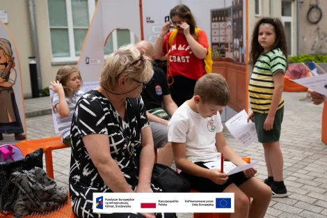 Grupa osób podczas otwarcia wystawy. Na pierwszym planie starsza kobieta w okularach i czarno-białej sukience wraz z chłopcem czytającym coś na trzymanej kartce. W tle inni zwiedzający wraz z dziećmi. 
