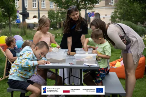 Grupa osób (dwie osoby dorosłe i czwórka dzieci) przy stole piknikowym podczas warsztatów archeologicznych. Jedna z dorosłych osób pochyla się nad jednym z dzieci przypatrując się jego twórczości. W tle plac Wolności oraz starsza kobieta w okularach przeciwsłonecznych siedząca na leżaku. 