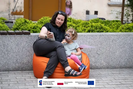 Ubrana na ciemno kobieta z okularami przeciwsłonecznymi założonymi jak opaska na włosy wraz z małą dziewczynką, która ma pomalowaną twarz we wzór kota siedzą na pomarańczowej ogromnej pufie i robią sobie zdjęcie selfie
