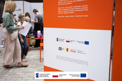 Na pierwszym planie jedna ze ścianek wystawy informująca o przyznanym dofinansowaniu z Unii Europejskiej oraz logotypami. W tle przechadzający się zwiedzający. 