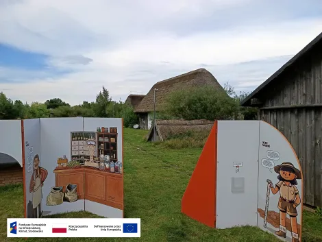 na pierwszym planie dwie ścianki wystawy mobilnej, na jednej z nich wnętrze sklepiku oraz wąsaty mężczyzna, na drugiej archeolożka. W tle zabudowania skansenu