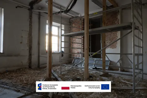 Wnętrze Muzeum Archeologicznego i Etnograficznego w Łodzi. Pomieszczenie w trakcie remontu z odsłoniętymi cegłami i fragmentami skutej ściany. Na środku stoi metalowe rusztowanie oraz pionowe drewniane belki konstrukcyjne, pod sufitem widoczne przewody instalacyjne. Na podłodze leży słoma i gruz budowlany, a z lewej strony światło wpada przez wysokie okna. Na dole grafika z logotypami Funduszy Europejskich, Rzeczypospolitej Polskiej oraz informacją o dofinansowaniu przez Unię Europejską.