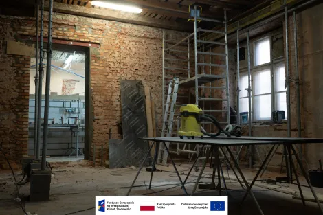 Wnętrze Muzeum Archeologicznego i Etnograficznego w Łodzi. Remontowane pomieszczenie z odsłoniętymi ceglanymi ścianami i drewnianym stropem. Na środku stoi stół roboczy z żółtym odkurzaczem budowlanym, wokół znajdują się rusztowania, drabina i narzędzia. Po prawej stronie widać wysokie okna, a po lewej przejście do kolejnego pomieszczenia w trakcie prac. Na dole grafika z logotypami Funduszy Europejskich, Rzeczypospolitej Polskiej i Unii Europejskiej.