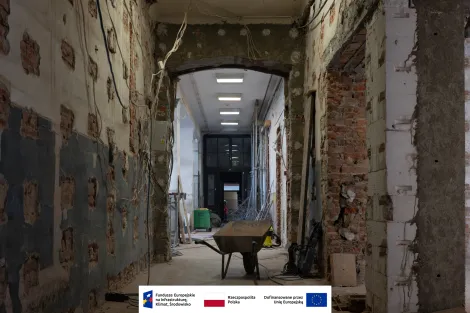 Wnętrze Muzeum Archeologicznego i Etnograficznego w Łodzi. Długi korytarz w trakcie remontu z odsłoniętą cegłą i fragmentami skutej farby na ścianach. Z sufitu zwisają przewody elektryczne, a na podłodze stoją taczka oraz sprzęt budowlany. W tle widać przeszklone drzwi i zieloną spawarkę na kółkach. Na dole obrazu znajduje się pasek z logotypami Funduszy Europejskich, Rzeczypospolitej Polskiej i Unii Europejskiej.