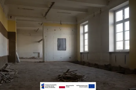 Wnętrze Muzeum Archeologicznego i Etnograficznego w Łodzi. Duże, puste pomieszczenie w trakcie remontu. Ściany z widocznymi otworami i szczelinami, fragmenty zerwanej podłogi oraz sterty drewnianych listew na ziemi. Po prawej stronie znajdują się wysokie okna wpuszczające światło dzienne. Na dole widoczne są logotypy Funduszy Europejskich, Rzeczypospolitej Polskiej oraz Unii Europejskiej