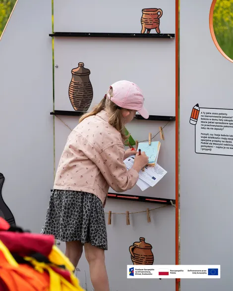 Dziecko w różowej czapce i jasnej kurtce zapisuje notatki przy interaktywnej tablicy edukacyjnej na świeżym powietrzu. Na tablicy znajdują się ilustracje glinianych naczyń oraz klamerki do przypinania kartek. Na dole zdjęcia znajduje się pasek z logotypami Funduszy Europejskich, Rzeczypospolitej Polskiej i Unii Europejskiej.