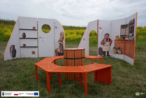 Plenerowa instalacja edukacyjna na trawiastym terenie. Wokół okrągłego stołu ustawiono pomarańczowe ławki oraz białe tablice z ilustracjami przedstawiającymi dawne naczynia, rzemieślniczkę i sklepikarza. W tle widoczne są kwitnące na żółto rośliny i pochmurne niebo. Na dole zdjęcia znajduje się pasek z logotypami Funduszy Europejskich, Rzeczypospolitej Polskiej i Unii Europejskiej.