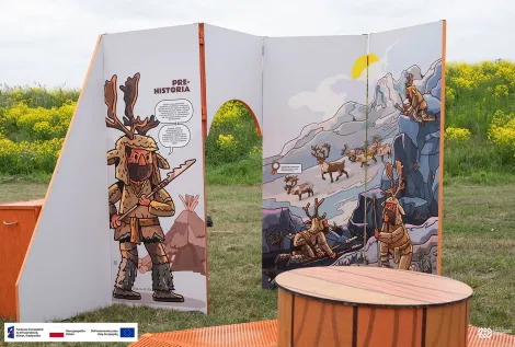 Fragment plenerowej instalacji edukacyjnej. Na białych tablicach widoczne są kolorowe ilustracje przedstawiające myśliwych ubranych w skóry zwierząt oraz stado jeleni w górskim krajobrazie. Na pierwszym planie znajduje się okrągły stół i pomarańczowa ławka, a w tle kwitnące na żółto rośliny. Na dole zdjęcia znajduje się pasek z logotypami Funduszy Europejskich, Rzeczypospolitej Polskiej i Unii Europejskiej.
