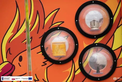 Zbliżenie na element interaktywnej instalacji edukacyjnej. Na pomarańczowym tle z ilustracją feniksa umieszczono trzy przezroczyste kopuły, wewnątrz których znajdują się eksponaty. Na dole zdjęcia znajduje się pasek z logotypami Funduszy Europejskich, Rzeczypospolitej Polskiej i Unii Europejskiej.