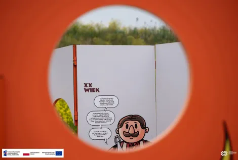 Widok przez okrągły otwór w pomarańczowej konstrukcji na planszę wystawową z rysunkową postacią i komiksowymi dymkami. Na dole zdjęcia znajduje się pasek z logotypami Funduszy Europejskich, Rzeczypospolitej Polskiej i Unii Europejskiej.