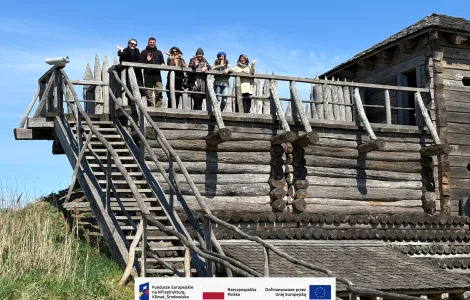 Sześć osób stoi na rekonstrukcji budowli z drewnianych bali i macha w stronę aparatu. Po prawej stronie widać fragment drewnianej wieży. W tle rozciąga się jasne, bezchmurne niebo, a wokół rośnie trawa. Na dole widoczne logotypy Rzeczypospolitej Polskiej oraz Funduszy Europejskich.