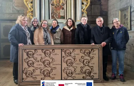 Grupa osób stoi we wnętrzu drewnianego kościoła przed ozdobionym ołtarzem z ikoną świętego. Wszyscy ustawieni są w jednym rzędzie i trzymają duży obraz na desce przedstawiający rzędy czaszek ze skrzyżowanymi kośćmi oraz łacińskie napisy, min. „Memento mori”. W tle widoczne są bogato zdobione elementy ołtarza oraz drewniane ściany świątyni. Na dole widoczne logotypy Rzeczypospolitej Polskiej oraz Funduszy Europejskich.
