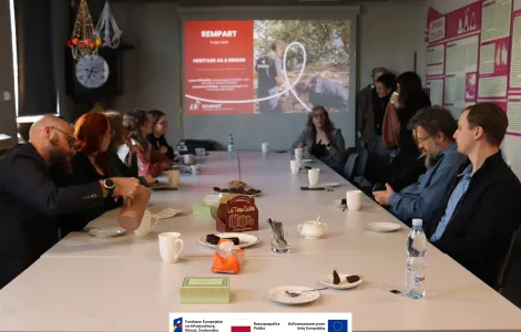 Grupa kilkunastu osób siedzi przy długim stole w sali konferencyjnej podczas spotkania. Na końcu stołu wyświetlany jest slajd prezentacji z tytułem „REMPART - Heritage as a Bridge”, a jedna z uczestniczek prowadzi dyskusję. Na stole stoją filiżanki, talerzyki z ciastkami i napojami. Uczestnicy słuchają i piją kawę, a na ścianach widać plansze informacyjne. Na dole widoczne logotypy Rzeczypospolitej Polskiej oraz Funduszy Europejskich.