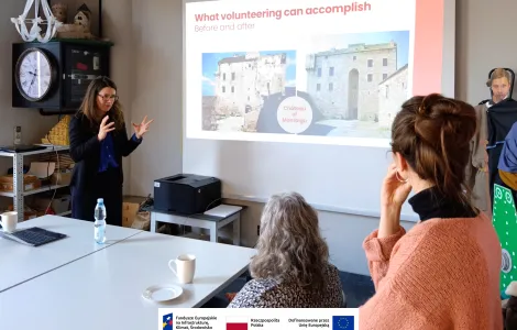 Kobieta prowadzi prezentację przed kilkoma słuchaczami w sali konferencyjnej. Stoi przy ekranie projekcyjnym i gestykuluje rękami, wyjaśniając temat. Na slajdzie widnieje tytuł „What volunteering can accomplish” oraz zdjęcia pokazujące efekt „przed i po” renowacji budynku. Uczestnicy siedzą przy stole, na którym stoją kubki, talerzyki i butelka wody, i uważnie słuchają prezentacji. Na dole widoczne logotypy Rzeczypospolitej Polskiej oraz Funduszy Europejskich.