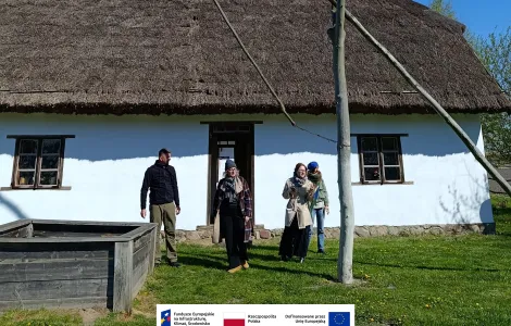 Cztery osoby stoją przed tradycyjną białą chatą z dachem krytym strzechą. Na pierwszym planie widać żuraw studzienny. Nad całą sceną rozciąga się bezchmurne, intensywnie niebieskie niebo, a wokół domu rośnie zielona trawa. Na dole widoczne logotypy Rzeczypospolitej Polskiej oraz Funduszy Europejskich.