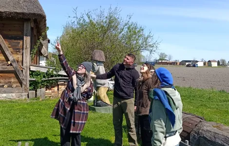 Cztery osoby stoją na trawie obok drewnianej chaty krytej strzechą. Jedna z nich wskazuje ręką w górę, a pozostali patrzą w tym samym kierunku. W tle widać krzewy, pole uprawne oraz kilka budynków, a nad całą sceną rozciąga się jasne, bezchmurne niebo. Na dole widoczne logotypy Rzeczypospolitej Polskiej oraz Funduszy Europejskich.