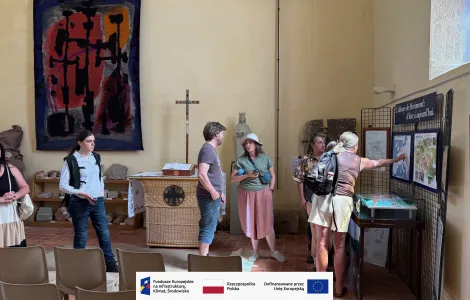 Grupa osób zwiedza wnętrze jasnego budynku. Kilka osób stoi przy tablicach informacyjnych i ogląda mapy oraz plansze wystawowe, jedna osoba wskazuje na jeden z elementów ekspozycji. W tle widoczny jest ołtarz z krzyżem, rzeźbą oraz tkaniną artystyczną zawieszoną na ścianie. W pomieszczeniu ustawione są także krzesła. Na dole obrazu znajduje się pasek z logotypami Funduszy Europejskich, Rzeczypospolitej Polskiej i Unii Europejskiej.