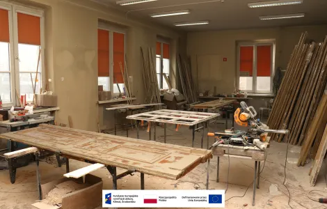 Wnętrze Muzeum Archeologicznego i Etnograficznego w Łodzi. Warsztat stolarski w trakcie renowacji drewnianych drzwi i okien. Na pierwszym planie leżą stare, szlifowane drzwi, obok stoją elektronarzędzia i stoły robocze. W tle widoczne są ramy okienne oraz elementy drewniane przygotowane do odnowienia. Pomieszczenie jest jasne, z dużymi oknami zasłoniętymi pomarańczowymi roletami. Na dole obrazu znajduje się pasek z logotypami Funduszy Europejskich, Rzeczypospolitej Polskiej i Unii Europejskiej.