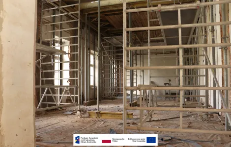 Pomieszczenie zabytkowego budynku podczas prac remontowych, wypełnione metalowymi rusztowaniami i podporami konstrukcyjnymi. Na podłodze widoczny jest pył budowlany oraz materiały zabezpieczające, a przez wysokie okna wpada światło dzienne oświetlające wnętrze. Na dole obrazu znajduje się pasek z logotypami Funduszy Europejskich, Rzeczypospolitej Polskiej i Unii Europejskiej.