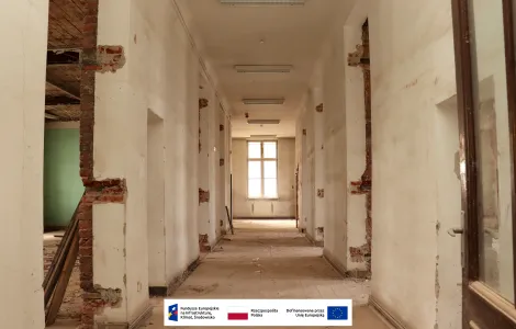 Długi korytarz zabytkowego budynku podczas prac remontowych. Częściowo odsłonięte ceglane ściany, zniszczone tynki i puste pomieszczenia po obu stronach. Na końcu korytarza widoczne wysokie okno wpuszczające światło dzienne. U dołu znajdują się logotypy Funduszy Europejskich, Rzeczypospolitej Polskiej i Unii Europejskiej.
