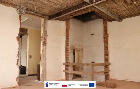 Pomieszczenie zabytkowego budynku w trakcie remontu. Częściowo rozebrane ściany odsłaniają ceglaną konstrukcję i drewniany strop ze stalowymi belkami. W centrum znajduje się zabezpieczony otwór w podłodze otoczony drewnianą barierką. U dołu widoczne są logotypy Funduszy Europejskich, Rzeczypospolitej Polskiej i Unii Europejskiej.