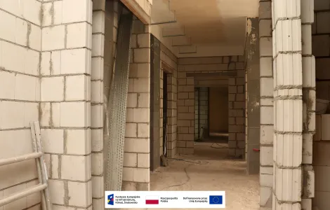 Korytarz w trakcie budowy wewnątrz budynku. Widoczne ściany z jasnych bloczków, nieotynkowane przejścia do pomieszczeń, sufit z elementami instalacji oraz materiały budowlane. Na dole grafiki znajdują się logotypy Funduszy Europejskich, Rzeczypospolitej Polskiej i Unii Europejskiej.