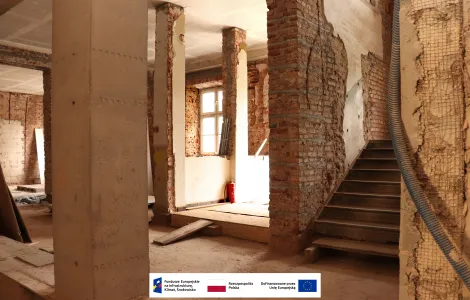 Wnętrze budynku podczas prac remontowych. Widoczne odsłonięte ceglane ściany, betonowe filary oraz schody prowadzące na wyższe piętro. Przez okna wpada światło dzienne, a w pomieszczeniu znajdują się elementy instalacji i materiały budowlane. Na dole zdjęcia umieszczono logotypy Funduszy Europejskich, Rzeczypospolitej Polskiej i Unii Europejskiej.