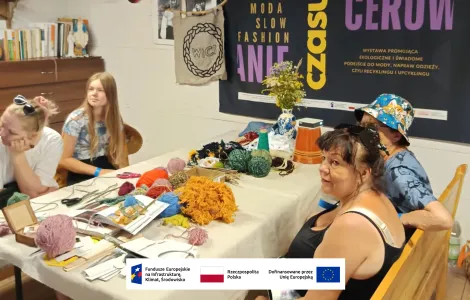 siedzące przy stole cztery kobiety - uczestniczki warsztatów, na stole leżą kolorowe włóczki, na ścianie wisi banner promujący wydarzenie. Kobiety patrzą w lewo (prawdopodobnie) na prowadzącą warsztaty