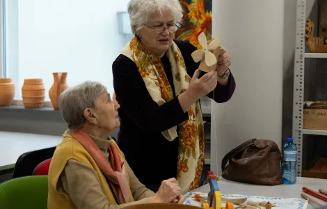 Starsza pani siedzi i patrzy na drewniany wiatraczek, który trzyma stojąca obok druga starsza pani. Na stole przed nimi leżą drewniane zabawki.