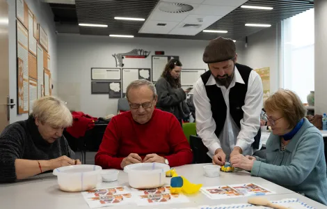 Troje starszych ludzi siedzi przy stole. Między nimi stoi mężczyzna w czapce. Ludzie coś związują. Przed nimi na stole leżą kolorowe kartki, łyżki i dwie miski.