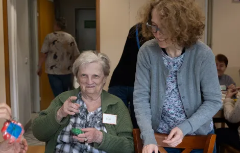 Starsza kobieta uśmiechnięta siedzi przy stole. Trzyma w ręku zieloną piłeczkę na gumce. Obok niej stoi uśmiechnięta druga kobieta.