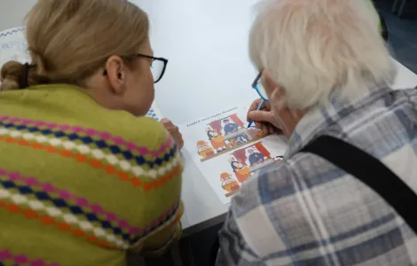 Przy stole widzimy tyłem starszego siwego mężczyznę i obok niego - kobietę. Patrzą na kartkę z kolorowym obrazkiem. Mężczyzna ma w ręku długopis.