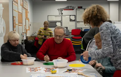 Grupa osób siedzi przy stole. Na stole leżą drewniane łyżki i dwie miski oraz kolorowe kartki. Obok stoi tyłem kobieta, która trzyma w ręku granatową taśmę.