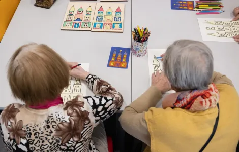 Dwie starsze panie kolorują obrazki. Między nimi leży wydrukowany kolorowy obrazek przedstawiający szopkę krakowską i stoi pudełko z kredkami.