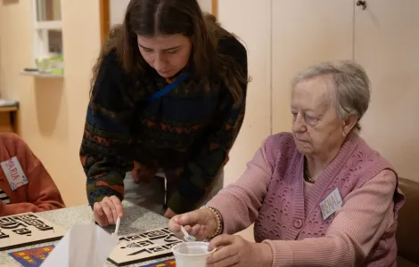 Starsza kobieta siedzi przy stole. Nabiera na małą łyżeczkę ryż z białego pojemnika. Obok niej stoi młoda kobieta z taką samą łyżeczką w ręku. Na stole leży drewniany szablon szopki krakowskiej.