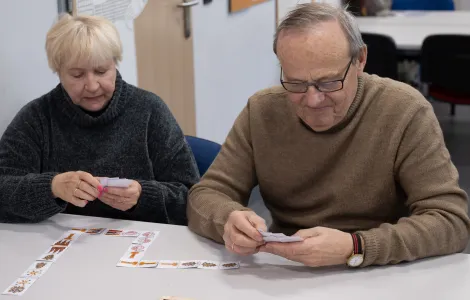 Kobieta i mężczyzna siedzą przy stole i układają domino. Są wpatrzeni w karteczki, które trzymają w dłoniach.