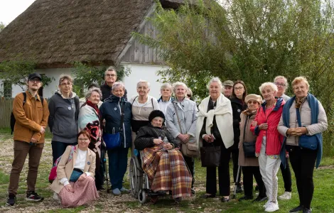 Na zdjęciu widoczna kilkunastoosobowa grupa, głównie starszych osób. Po środku nich na wózku siedzi starsza kobieta owinięta kocem. Obok niej kuca młoda kobieta. Reszta osób stoi. 