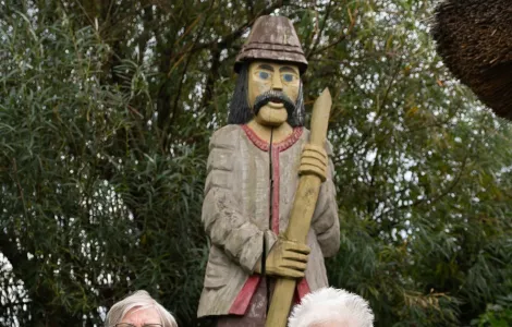 Dwie starsze kobiety w kurtkach usiadły na krzesełkach turystycznych. Za nimi widać dużą drewnianą rzeźbę przedstawiającą mężczyznę.