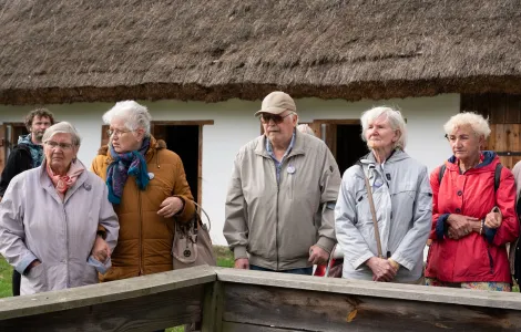 Na pierwszym planie stoi grupa pięciu seniorów ubranych w kurtki. Za nimi widać mężczyznę z brodą. W tle dach wiejska chałupa kryta słomą.