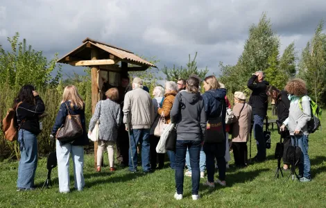 Grupa osób w kurtkach stoi i patrzy na drewnianą tablicę przykrytą daszkiem. Obok tablicy stoi  mężczyzna i coś pokazuje ręką. Z tyłu widać drzewa i trawy.