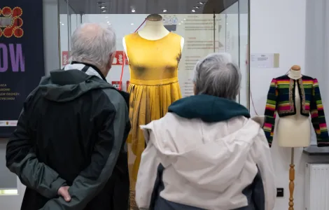 Dwoje starszych ludzi wpatruje się w gablotę, w której umieszczono żółtą sukienkę.