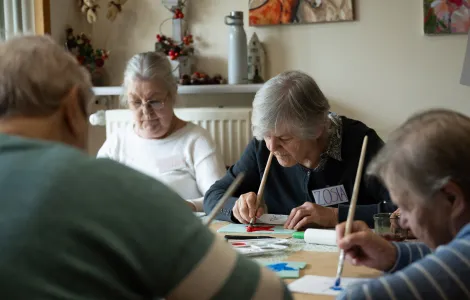Cztery starsze osoby siedzą przy stole. Malują farbami coś na kartce.