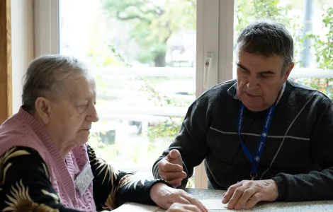 Dwoje starszych ludzi siedzi przy stole. Patrzą na leżącą na stole planszę z wiejską chałupą.