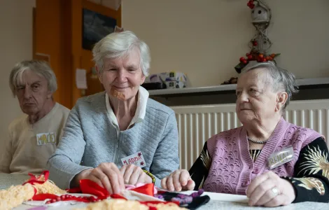 Dwie starsze kobiety i starszy mężczyzna siedzą przy stole. Kobieta siedząca po środku dotyka planszy wykonanej z materiału i włóczki.