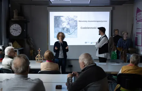 Grupa ludzi patrzy na ekran z napisem "Warsztaty muzeoterapeutyczne. Codzienność". Przed ekranem stoi mężczyzna w czapce z daszkiem i kobieta z kręconymi włosami.