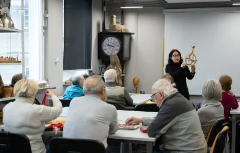 Grupa ludzi siedzi przy stolikach i patrzy na kobietę ubraną na czarno. kobieta trzyma w ręku wieniec dożynkowy wykonany z wikliny.