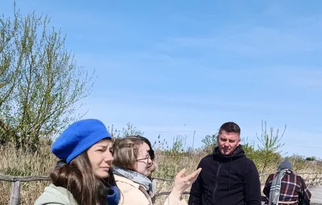 Grupa czterech osób stoi na drewnianej kładce na świeżym powietrzu i ogląda tablicę informacyjną. Dwie kobiety na pierwszym planie  (jedna w niebieskim berecie i zielonej kurtce, druga w jasnym płaszczu i okularach) rozmawiają, a jedna z nich gestykuluje ręką w stronę tablicy. Obok stoi mężczyzna w czarnej bluzie, patrzący na planszę. W tle widać roślinność, drewniane ogrodzenie i bezchmurne niebo. Na dole widoczne logotypy Rzeczypospolitej Polskiej oraz Funduszy Europejskich.